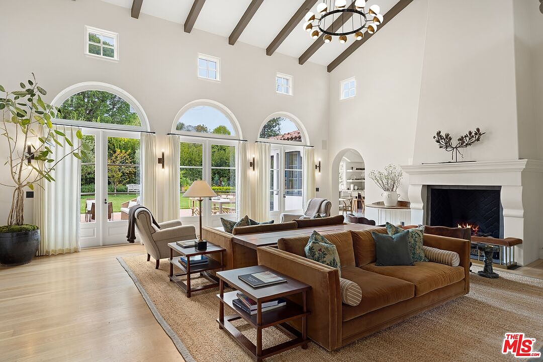 This is a spacious living room with high ceilings, exposed beams, and large arched windows that provide ample natural light and views of the exterior. The room features a comfortable brown sofa, a fireplace, and elegant decor, creating a warm and inviting atmosphere. The perspective is from the front, showcasing the room's depth and architectural details.