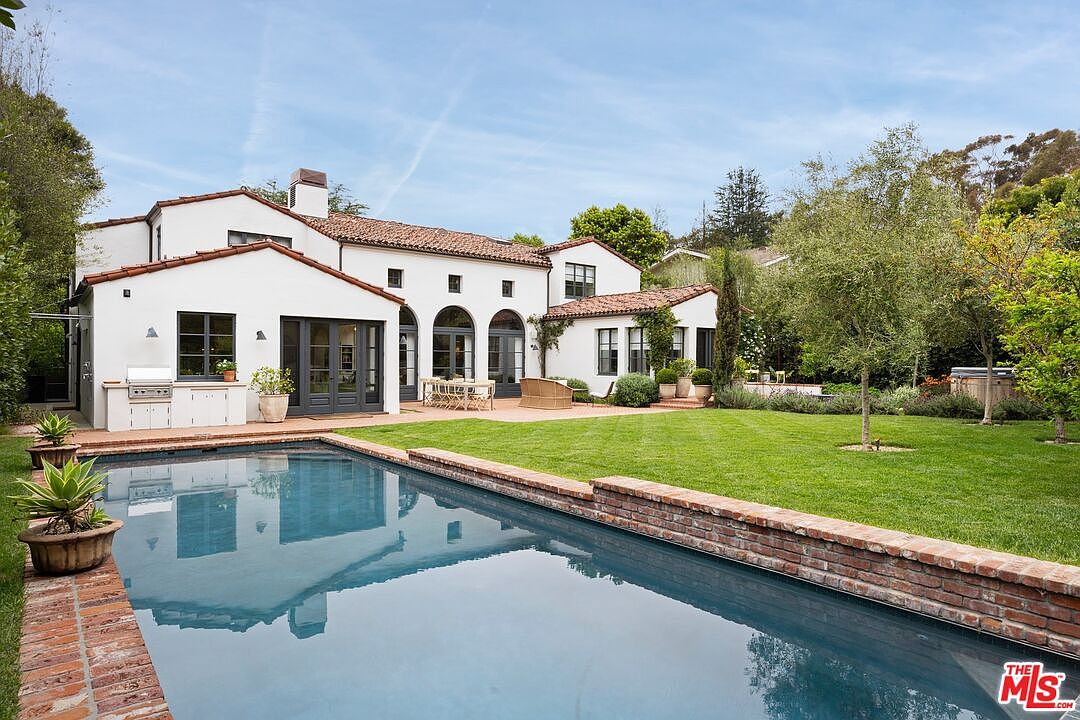 This image showcases the rear exterior of a luxurious home, highlighting a stunning pool area. The pool, with its brick border, reflects the house's white facade and terracotta roof tiles. A well-manicured lawn extends from the pool area, leading to mature trees and landscaping, suggesting a private and serene backyard oasis.