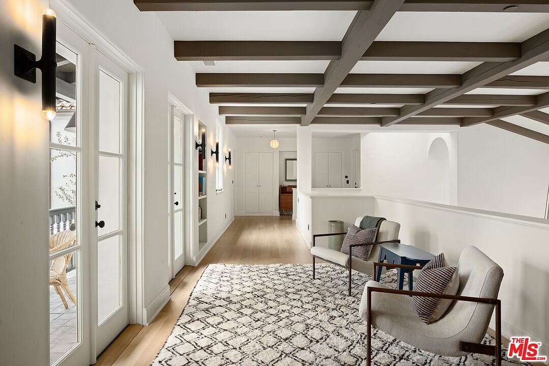 This interior shot showcases a bright hallway with white walls, light wood flooring, and a striking coffered ceiling with dark wood beams. The hallway features French doors leading to an outdoor space, built-in shelving, and stylish sconce lighting. Two armchairs and a patterned rug create a cozy seating area at the end of the hallway, adding a touch of elegance and comfort.
