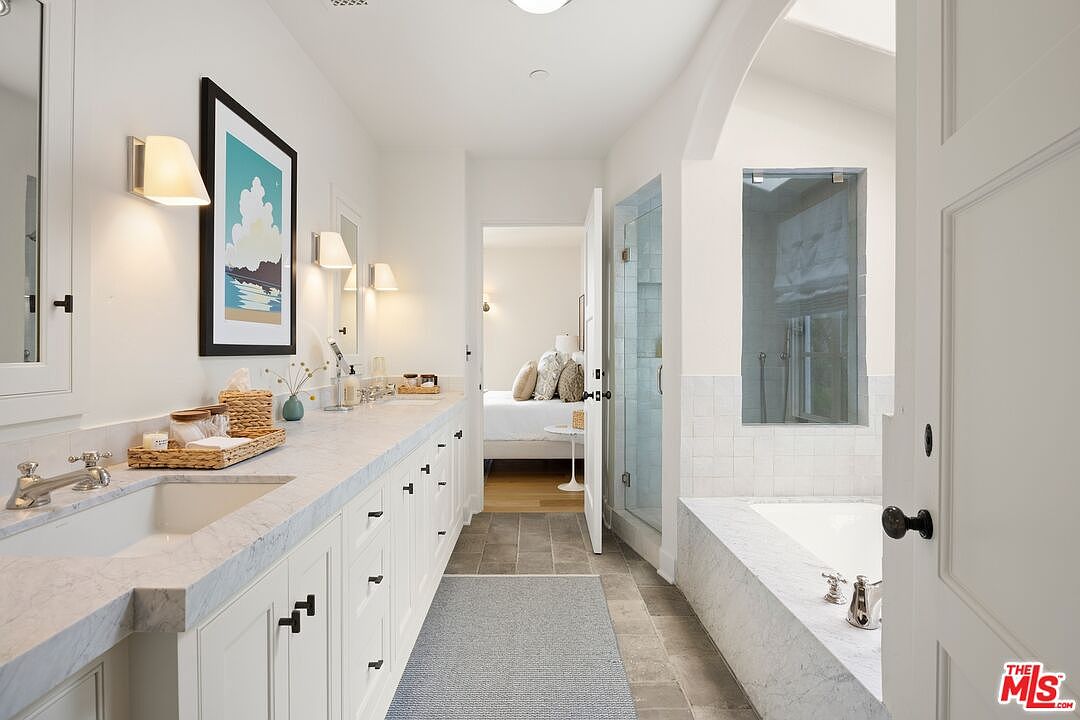 This is a bright and spacious primary bathroom featuring a long vanity with marble countertops and white cabinetry. The room includes a walk-in shower with glass doors and a separate bathtub, also surrounded by marble. The color palette is predominantly white, creating a clean and luxurious atmosphere, with a doorway leading to the bedroom.