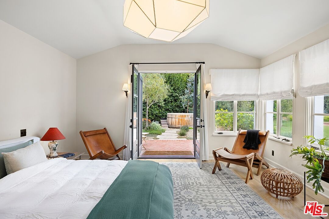 This is a bright and airy primary bedroom featuring a king-size bed with white linens and a teal throw. French doors open to a private patio with a hot tub, and large windows with white roman shades provide ample natural light. The room is decorated with neutral tones and natural materials, creating a relaxing and inviting atmosphere.