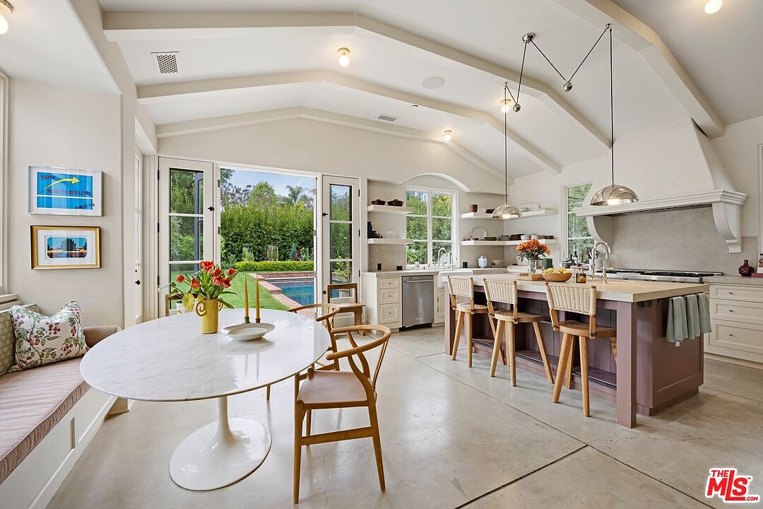 This is a bright and airy kitchen featuring a large marble-topped dining table with seating, a central island with bar stools, and a view of the pool through large glass doors. The kitchen is well-lit with a combination of natural light and pendant lighting, and the neutral color palette creates a clean and modern aesthetic. The open layout seamlessly connects the dining area with the kitchen space, making it ideal for entertaining.