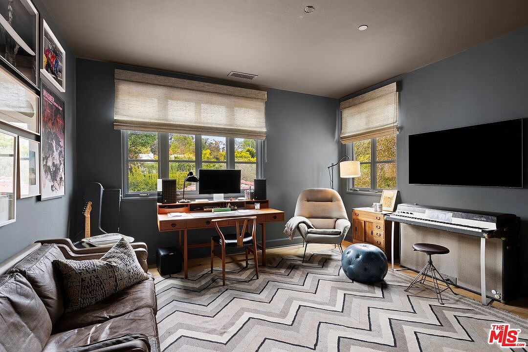 This is an interior shot of a home office or study. The room features a desk with a computer setup, a comfortable chair, a piano, and a leather sofa, suggesting a space for both work and creative pursuits. The chevron patterned rug adds a stylish touch to the room.