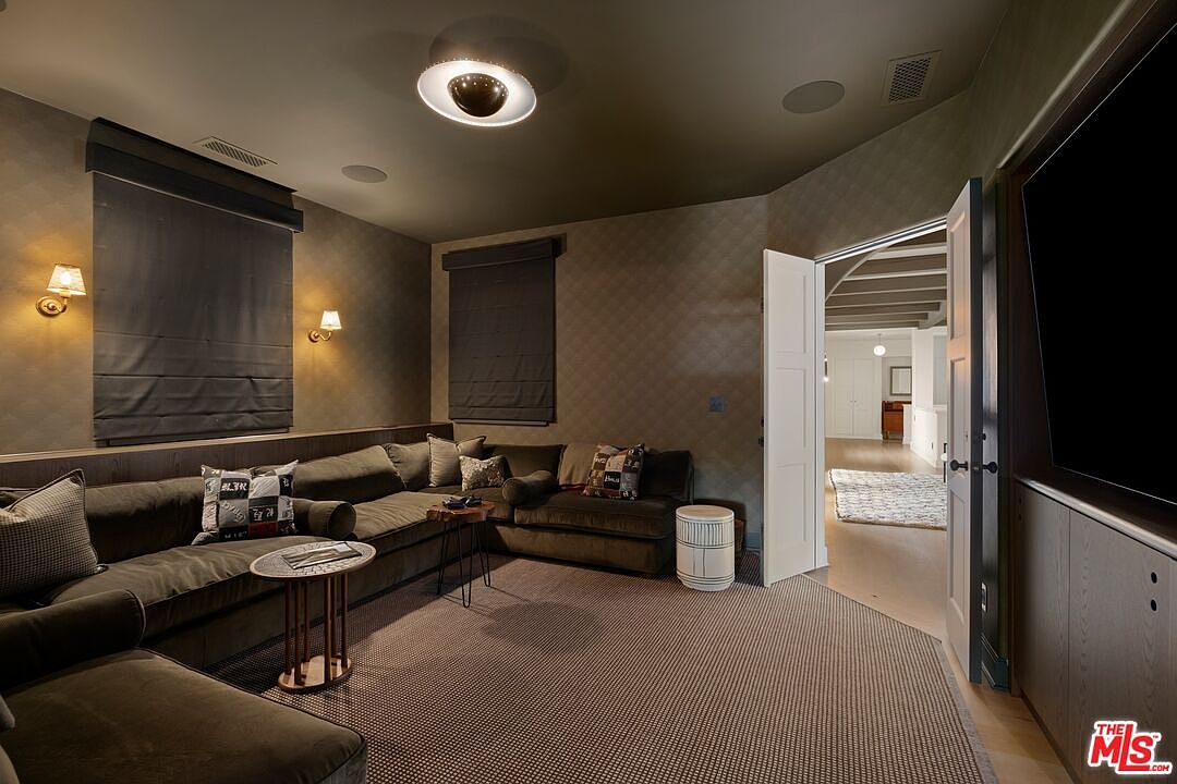 This is an interior shot of a cozy living room featuring a large, comfortable sectional sofa with decorative pillows. The room is illuminated by wall sconces and a modern ceiling light, creating a warm ambiance. The walls are covered in a subtle patterned wallpaper, and the flooring is a textured carpet. The perspective is from a low angle, emphasizing the depth and comfort of the space.