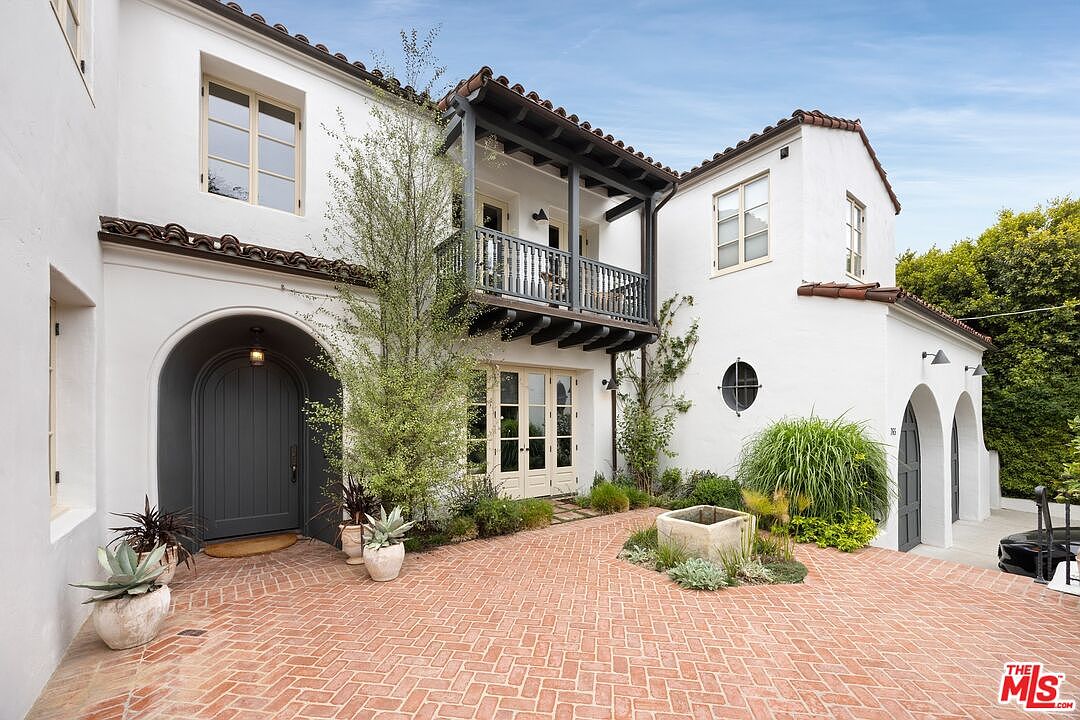 This is a front exterior view of a two-story Spanish-style home with a red brick courtyard. The house features white stucco walls, a red tile roof, arched doorways, and a balcony with dark railings. Lush landscaping and a decorative water feature add to the property's curb appeal.