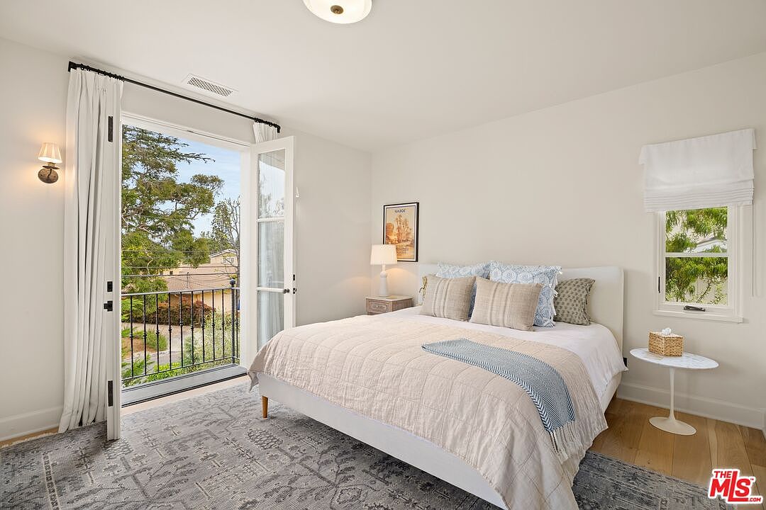 This is a bright and airy primary bedroom featuring a bed with neutral-toned bedding and pillows, a small side table, and a window with a white shade. The room has a balcony accessible through French doors, offering an indoor-outdoor feel. The walls are painted in a light neutral color, and a patterned rug covers the hardwood floor.
