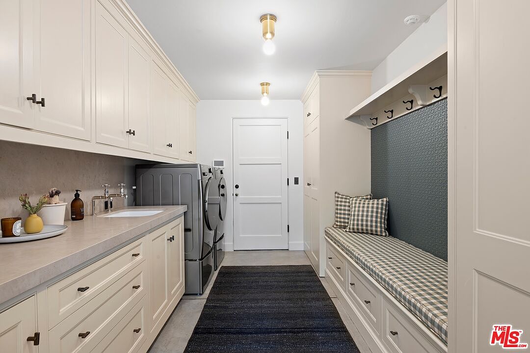 This is a well-organized laundry room featuring built-in cabinetry in a light neutral tone, providing ample storage space. A front-loading washer and dryer are neatly installed, and a long countertop with a sink offers a practical workspace. The room also includes a built-in bench with storage drawers and coat hooks, enhancing its functionality and appeal.