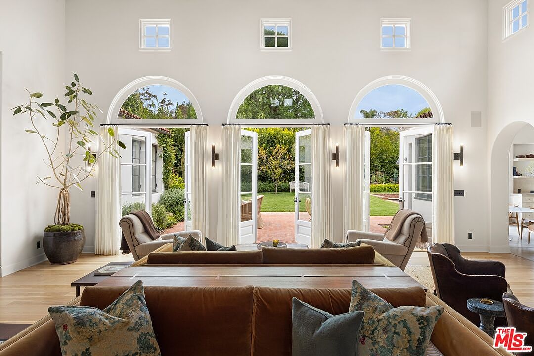 This is an interior shot of a living room featuring three arched doorways leading to a garden. The room is decorated with neutral tones, including a large brown sofa with decorative pillows, armchairs, and a tall potted plant. The high ceilings and natural light create an open and airy atmosphere, perfect for relaxation and entertaining.