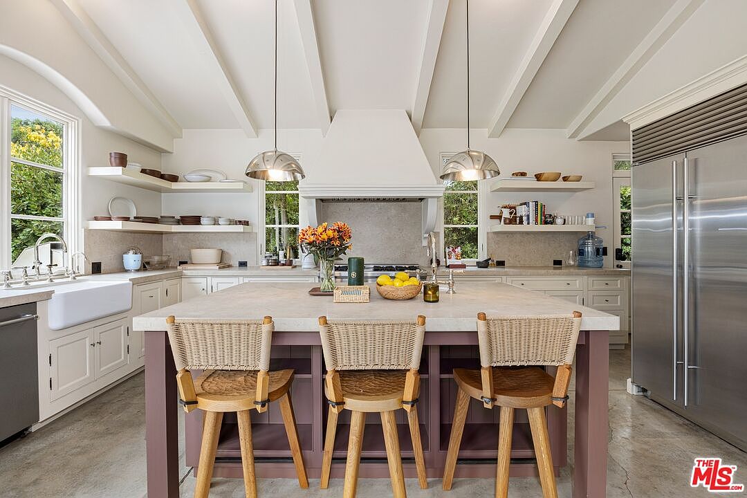 This is a bright and airy kitchen featuring a large center island with seating, custom cabinetry, and stainless steel appliances. The kitchen is well-lit with pendant lighting and natural light from the windows, creating a warm and inviting atmosphere. The perspective is from the front, showcasing the island and the overall layout of the kitchen.