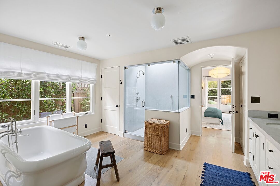This is a bright and airy primary bathroom featuring a freestanding bathtub positioned near a large window with white blinds. A glass-enclosed shower is adjacent to a woven basket, and a wooden stool sits on a gray rug. The room has light hardwood floors and leads to a bedroom through an arched doorway, creating a spa-like and luxurious atmosphere.