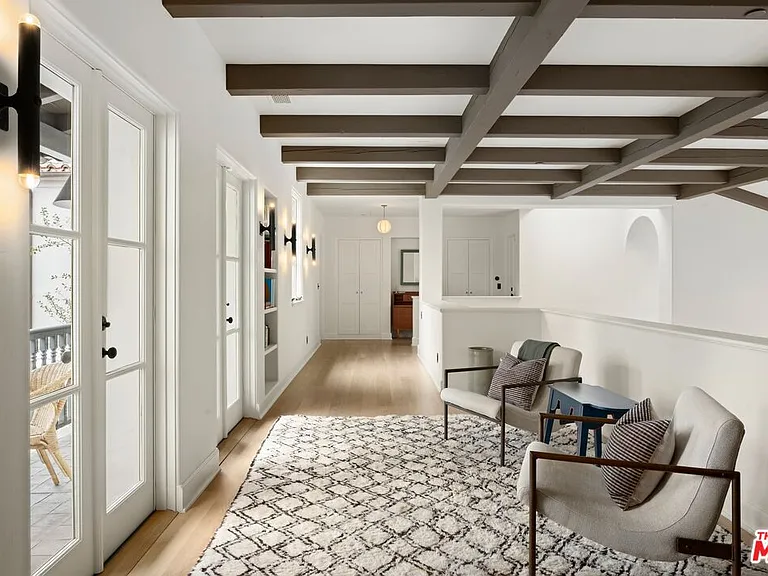 This interior shot showcases a bright and airy hallway on an upper level. Key features include exposed wood beam ceilings, white walls, light hardwood flooring, and a textured area rug underneath two accent chairs with a small table. The space gives a sense of luxury and modern design, with access to an outdoor balcony through glass doors.