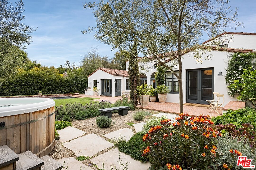 This image showcases a beautifully landscaped backyard featuring a wooden hot tub, stone pathways, and vibrant orange flowers. The white stucco house with dark-framed windows and a red tile roof provides a charming backdrop. The scene evokes a sense of tranquility and outdoor living.