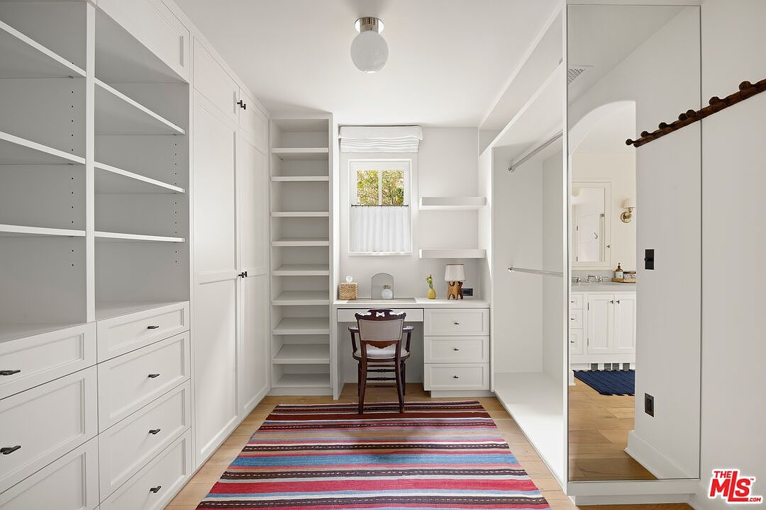 This is an interior shot of a well-organized walk-in closet. The closet features custom white cabinetry with drawers and shelving, a built-in desk area with a chair, and a colorful striped rug on the hardwood floor. A mirrored door provides a glimpse into an adjacent bathroom, enhancing the sense of space and luxury.