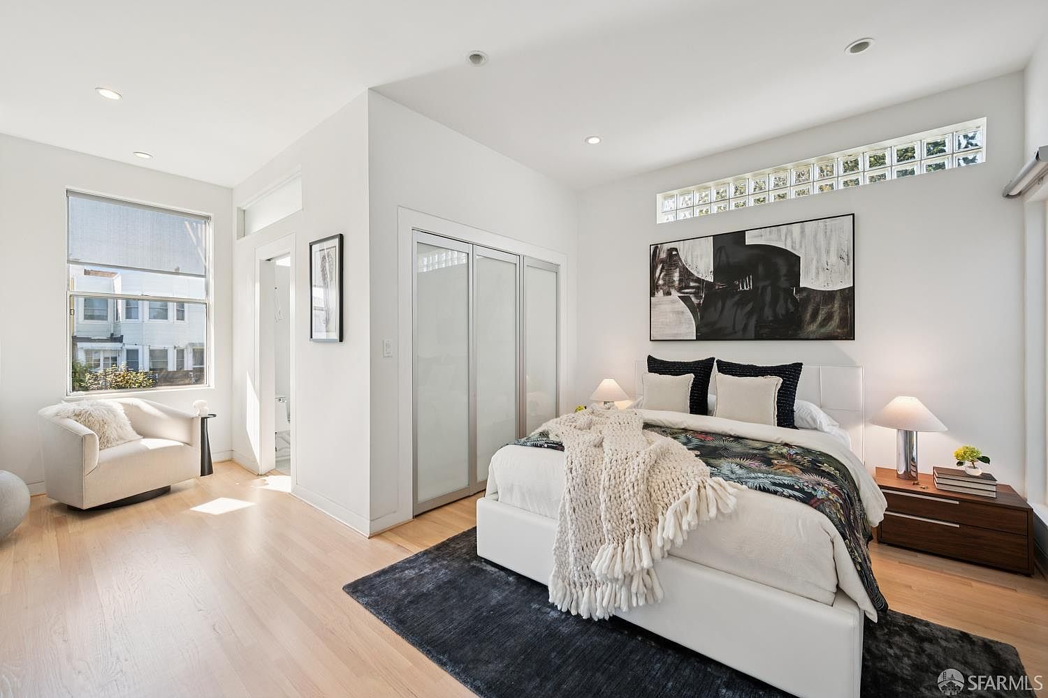 This bright and modern primary bedroom features light wood flooring, a plush white bed with a textured throw, and a dark area rug that anchors the space. The room includes a cozy armchair by the window, a sleek closet with frosted glass doors, and a unique horizontal row of glass block windows above the bed. The minimalist aesthetic is complemented by contemporary bedside lamps and a large abstract art piece, creating a serene and sophisticated atmosphere.