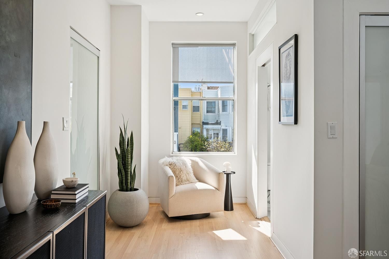 This bright and airy nook serves as a peaceful reading area, featuring a comfortable cream-colored swivel chair accented with a plush throw pillow. A tall, slender snake plant in a rounded concrete planter adds a touch of greenery, while a dark wood console table provides a sleek surface for decorative vases. The space is illuminated by a large window that frames a view of neighboring buildings, creating a serene and inviting atmosphere.