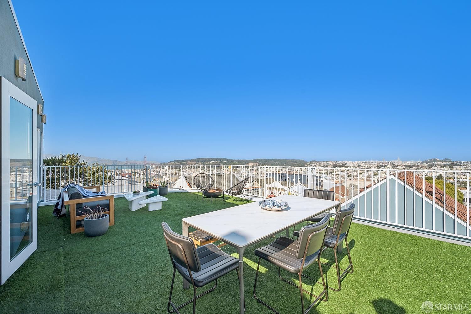 This expansive rooftop deck features artificial turf flooring and offers panoramic views of the city skyline and the Golden Gate Bridge. The space is furnished with a modern dining set and casual lounge seating, creating an ideal environment for outdoor entertaining. A white metal railing surrounds the perimeter, ensuring safety while maintaining the open, airy feel of this elevated outdoor living area.