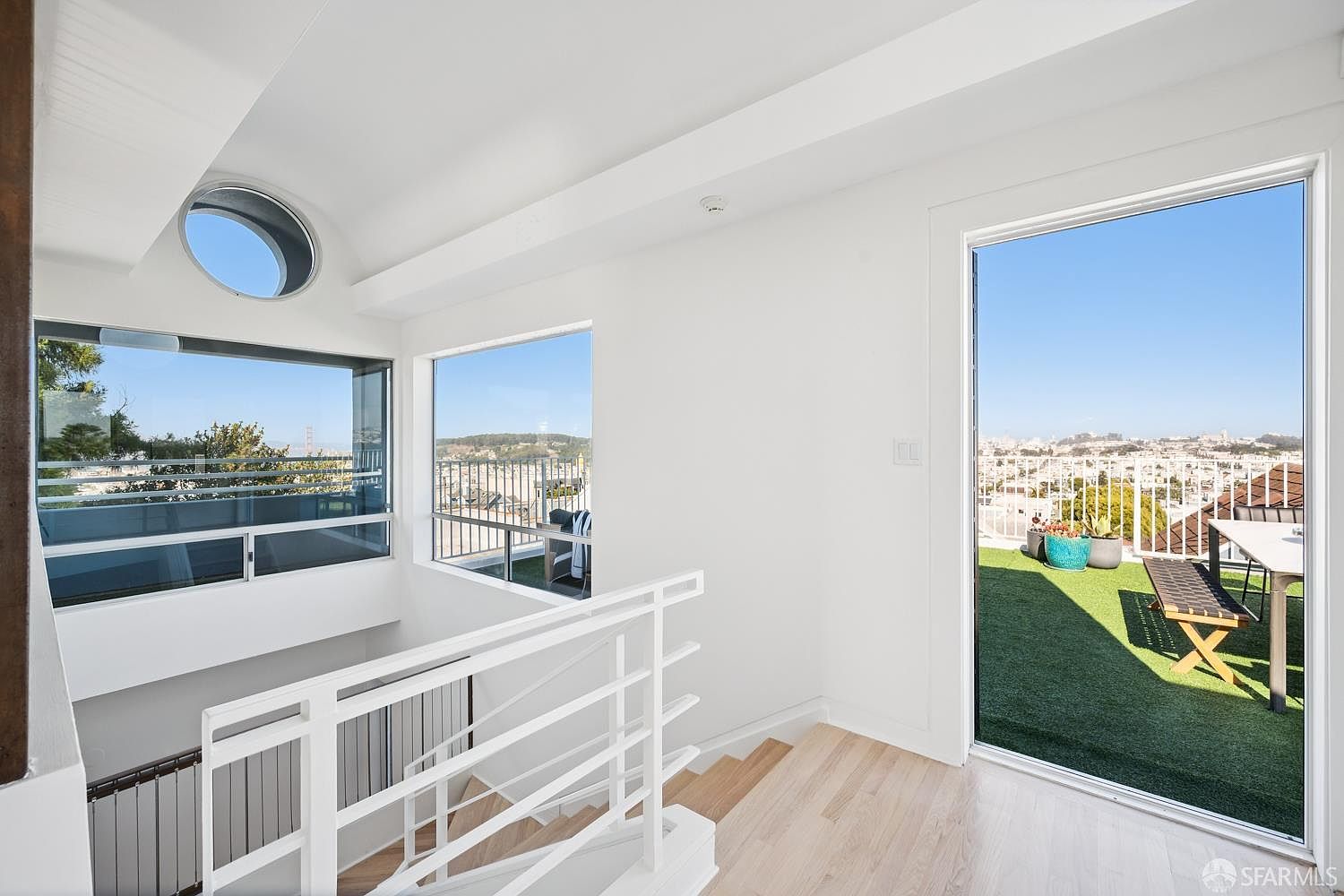 This bright, modern landing area features light wood flooring and a crisp white color palette, creating an airy and open atmosphere. A large glass door provides seamless access to an outdoor deck, while a unique circular window and large rectangular windows offer expansive views of the surrounding cityscape. The space is defined by clean lines, a minimalist white staircase railing, and a sense of architectural sophistication.