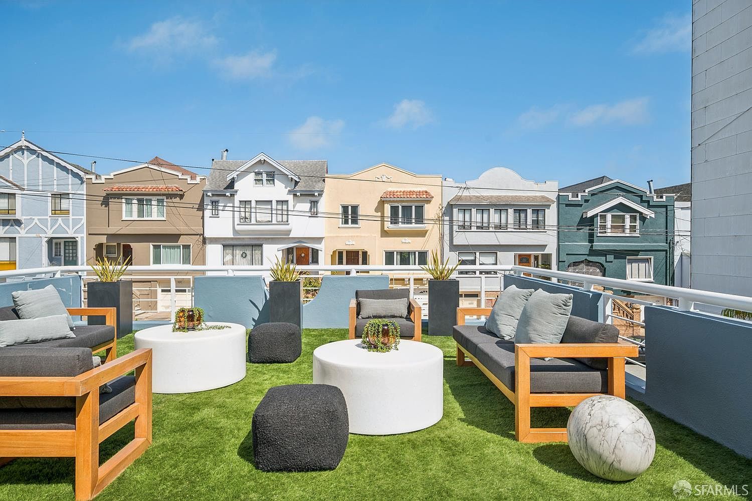 This rooftop deck offers a modern outdoor living space with artificial turf, contemporary wooden-framed lounge seating, and circular white tables. The area is framed by a sleek metal railing and provides an elevated perspective overlooking a row of colorful, classic San Francisco-style residential buildings. The bright, sunny atmosphere creates an inviting space for relaxation or entertaining.