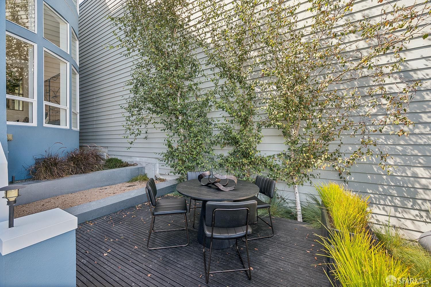 This outdoor patio features a dark-stained wooden deck furnished with a modern circular table and four sleek, black metal chairs. The space is framed by a blue-painted exterior wall with tall windows and a light-colored horizontal siding wall adorned with two slender birch trees. A tiered concrete planter box and a low-profile garden bed with ornamental grasses add texture and greenery to this intimate, contemporary outdoor living area.