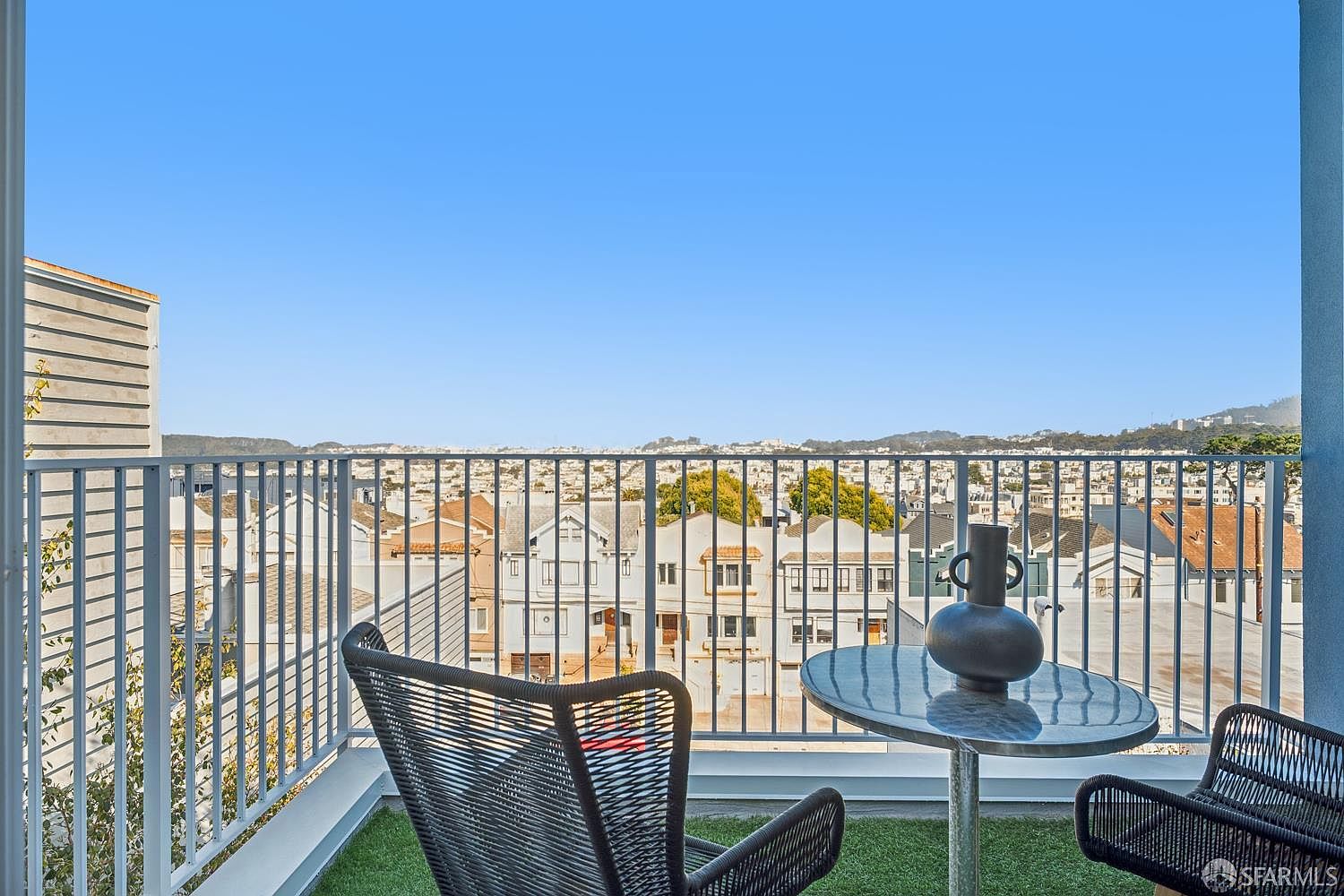 This elevated balcony offers a panoramic view of a dense urban neighborhood under a clear blue sky. The space is furnished with two modern, woven-style chairs and a small circular table topped with a decorative dark vase. The balcony features artificial turf flooring and a clean, white metal railing, creating a bright and inviting outdoor retreat.