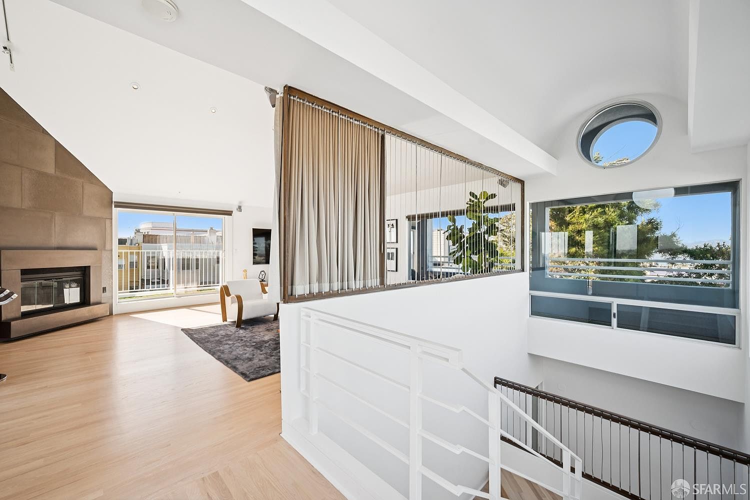 This bright, contemporary living space features light wood flooring and a prominent stone-clad chimney breast housing a fireplace. A unique architectural partition with vertical metal rods separates the living area from the staircase, which leads down to a lower level. Large windows and a circular accent window allow for abundant natural light, creating an airy and open atmosphere.