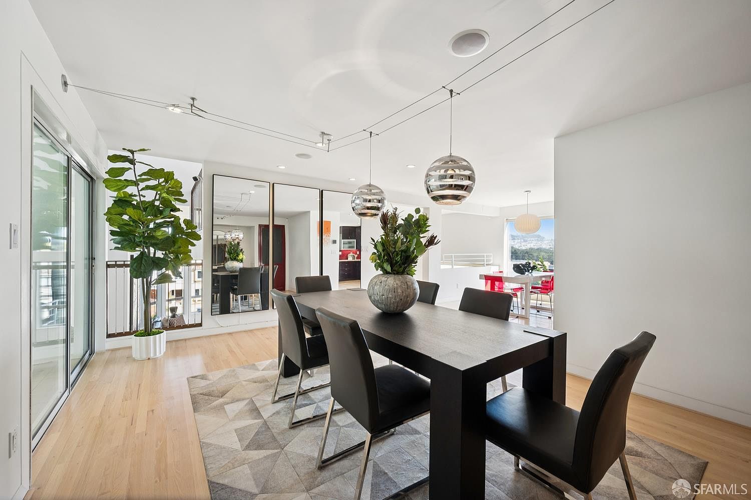 This contemporary dining room features a dark rectangular table surrounded by six sleek black chairs, all set atop a geometric-patterned area rug. The space is illuminated by two modern spherical pendant lights and includes a large potted plant near the sliding glass doors, which provide access to an outdoor area. A series of tall mirrors on the wall reflects the adjacent kitchen space, creating an open and airy atmosphere with light wood flooring throughout.