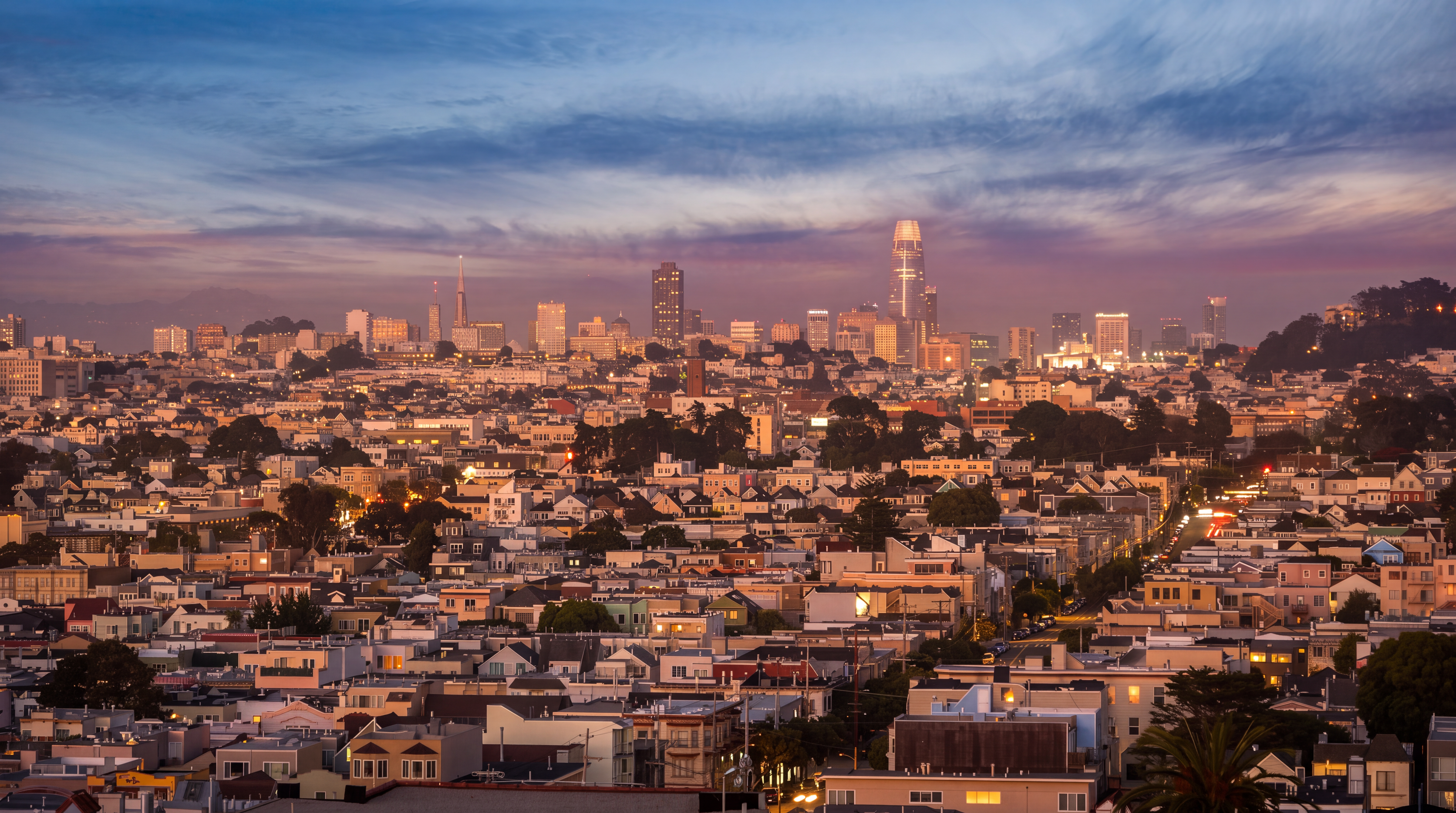 This wide-angle aerial view captures a sprawling cityscape at dusk, showcasing a dense urban landscape of residential buildings and high-rise towers. The iconic Salesforce Tower stands prominently on the horizon against a soft, gradient sky transitioning from deep blue to a warm, hazy sunset glow. The perspective provides a comprehensive look at the neighborhood's density and the surrounding metropolitan skyline.
