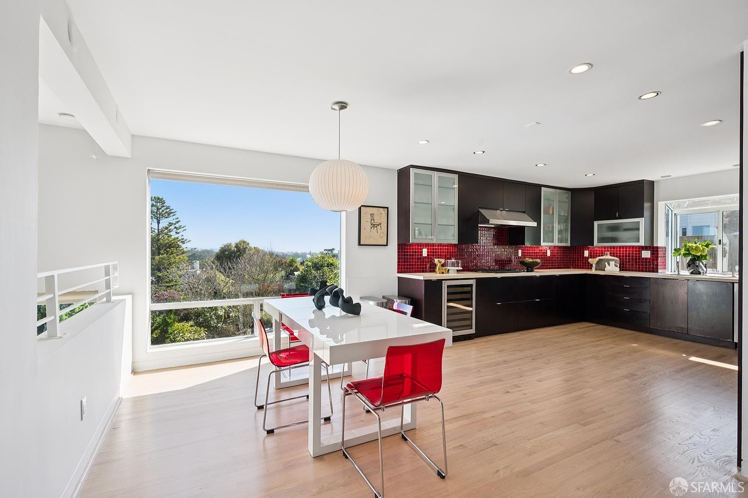 This open-concept kitchen and dining area features dark cabinetry contrasted by a vibrant red tiled backsplash and light wood flooring. A modern white dining table with red transparent chairs sits beneath a spherical pendant light, positioned next to a large window that provides an expansive view of the outdoors. The space is bright and contemporary, with recessed lighting and a wine refrigerator integrated into the cabinetry.