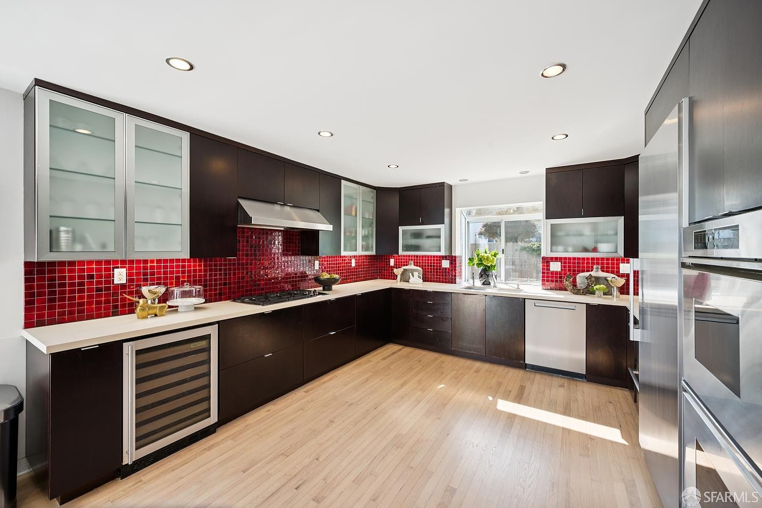 This modern kitchen features dark cabinetry contrasted by a vibrant red tiled backsplash and light-toned wood flooring. The space is equipped with stainless steel appliances, including a built-in wine cooler, a gas cooktop, and a wall-mounted oven. A window above the sink provides natural light, creating an open and functional layout.