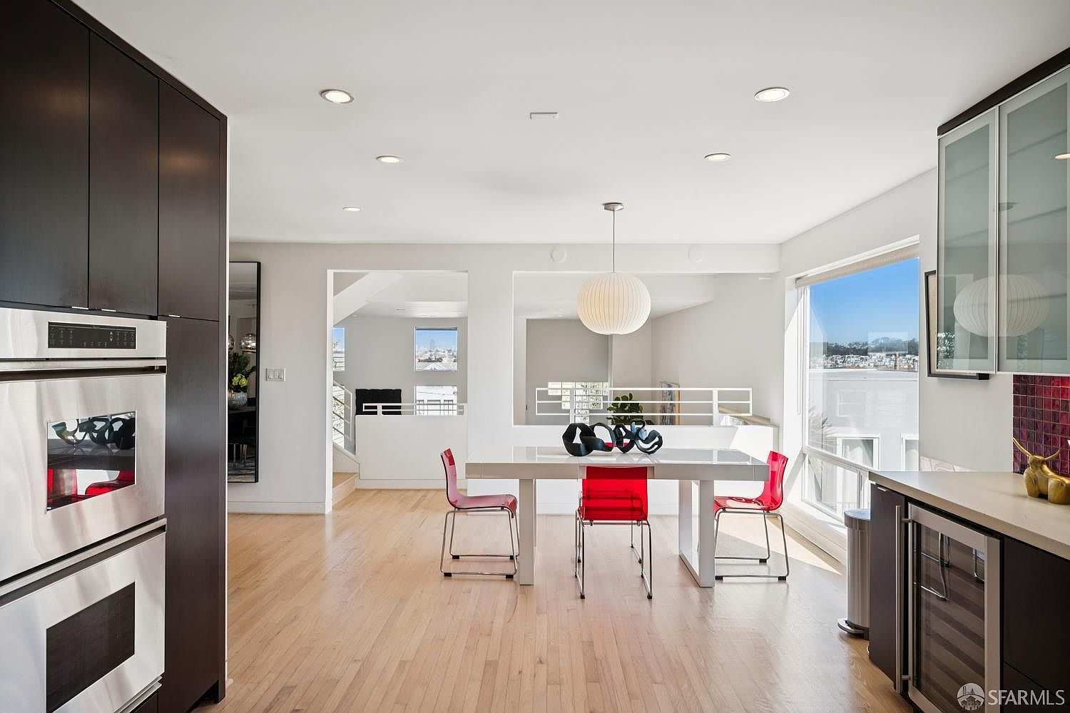 This bright and open dining area features a sleek white table paired with three vibrant red chairs, illuminated by a modern spherical pendant light. The space flows seamlessly into an adjacent living area, with light wood flooring throughout and large windows that provide ample natural light and a view of the neighborhood. The kitchen, visible on the left, includes dark cabinetry and stainless steel appliances, creating a contemporary and airy atmosphere.