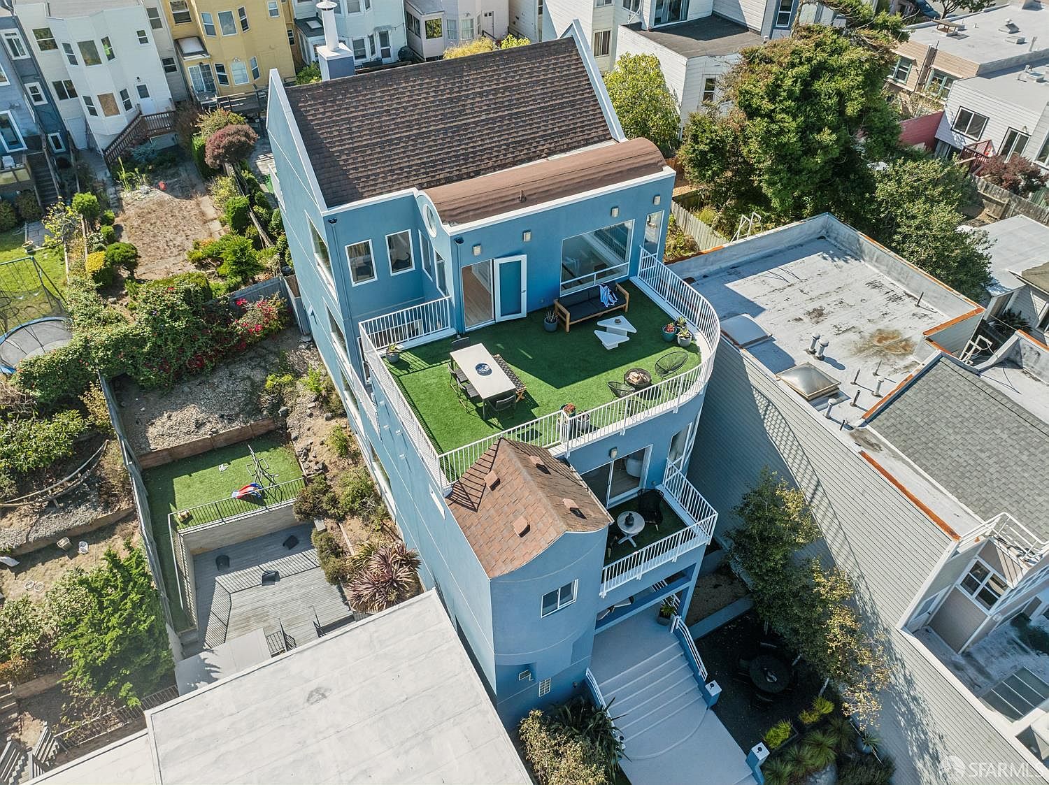 This high-angle aerial view captures a multi-level blue residential home featuring a prominent rooftop deck with artificial turf and outdoor dining furniture. The property includes a tiered backyard with a wooden deck and a lower-level patio, showcasing a blend of urban living and private outdoor space. The perspective highlights the home's architectural layout, including its curved balcony and the surrounding neighborhood density.