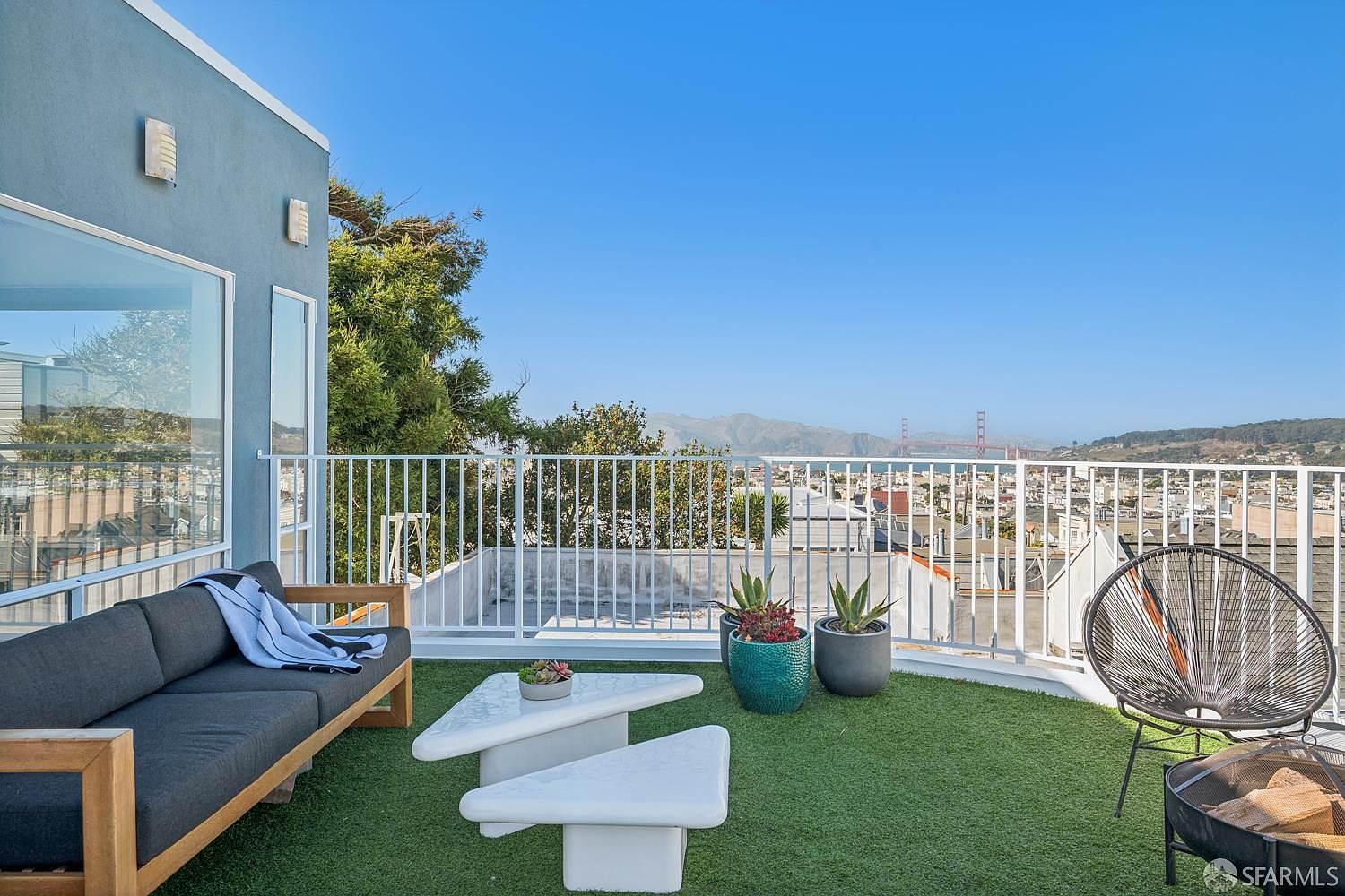 This elevated outdoor terrace features artificial turf flooring and a modern seating area with a dark gray sofa and a unique, tiered white coffee table. A sleek metal railing borders the space, providing an unobstructed, panoramic view of the city skyline and the iconic Golden Gate Bridge in the distance. The scene is captured from a slightly elevated perspective, emphasizing the seamless blend of contemporary outdoor living and a dramatic urban backdrop.
