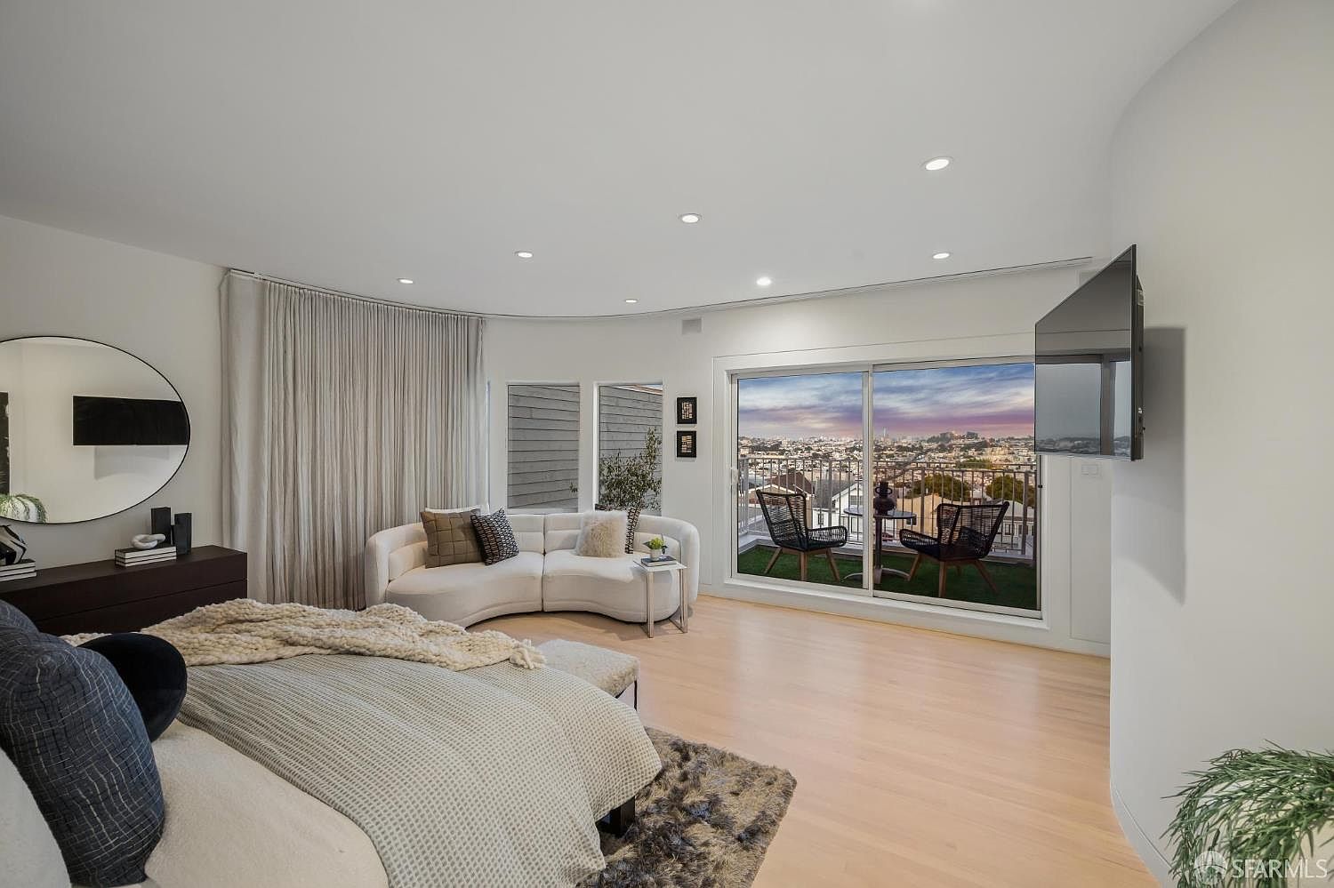 This spacious primary bedroom features light wood flooring, a curved white sofa, and a large circular mirror mounted on the wall. A sliding glass door provides access to a private balcony with outdoor seating and a panoramic city view. The room is illuminated by recessed lighting and offers a bright, contemporary atmosphere.