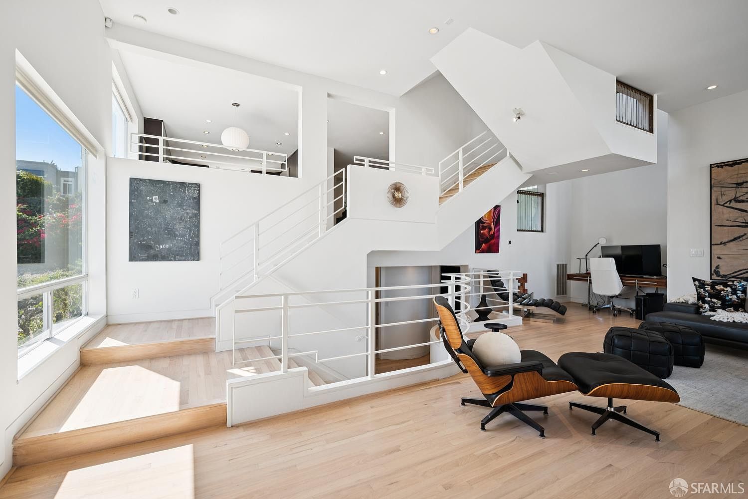 This bright, multi-level living space features a modern open-concept design with light wood flooring and a striking white staircase as a central architectural element. The room includes a comfortable lounge area with a classic mid-century modern chair and ottoman, a dedicated workspace with a desk, and large windows that flood the area with natural light. The clean, minimalist aesthetic is complemented by bold abstract artwork and a split-level layout that creates distinct zones within the open floor plan.