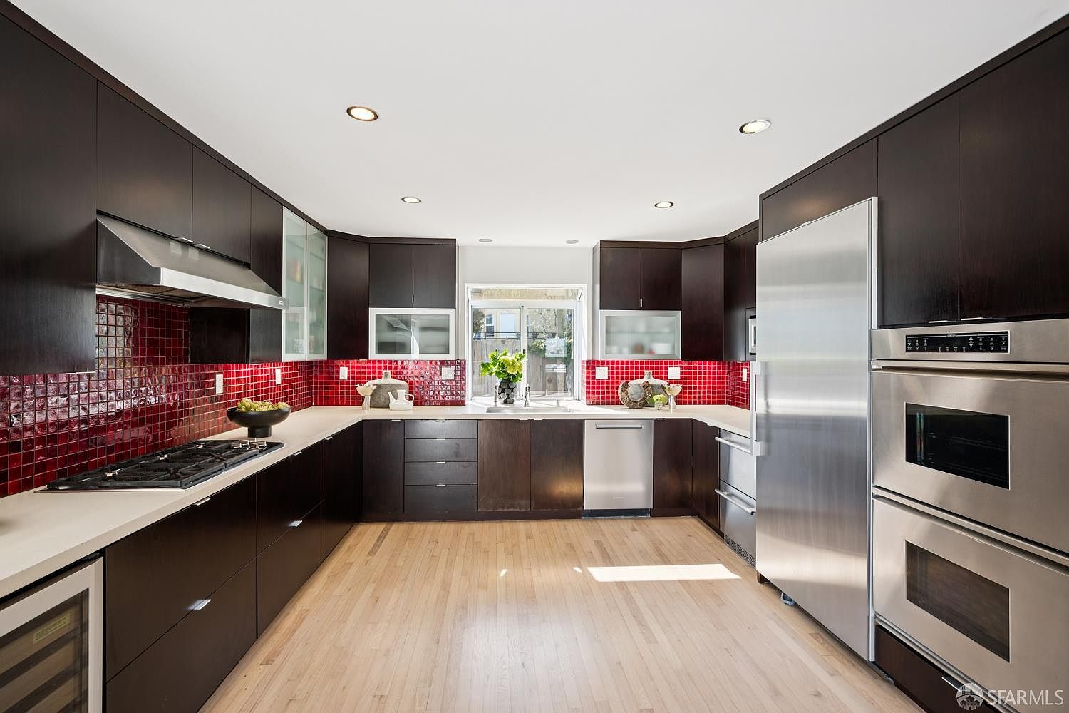 This modern kitchen features dark, flat-panel cabinetry contrasted by a vibrant red tiled backsplash and light-toned wood flooring. The space is equipped with stainless steel appliances, including a double wall oven and a large refrigerator, arranged in a functional U-shaped layout. A central window above the sink brings in natural light, creating an open and airy atmosphere in this contemporary culinary space.