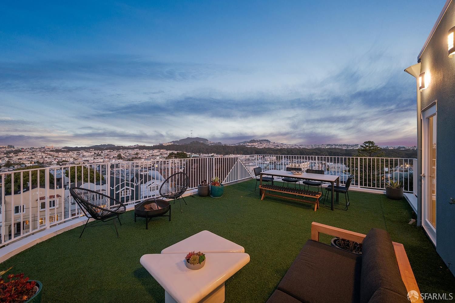 This expansive rooftop deck offers a panoramic view of the city skyline under a soft twilight sky. The space is furnished with a modern outdoor dining set, a comfortable lounge area with a dark sofa, and a fire pit, all set atop artificial turf. The elevated perspective highlights the open-air layout, making it an ideal setting for evening entertaining.