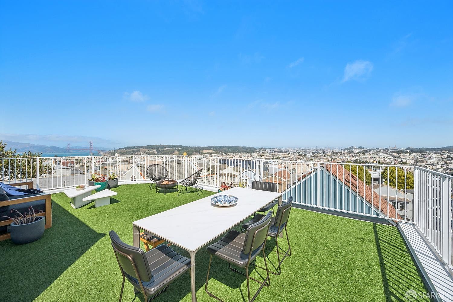 This expansive rooftop deck offers a panoramic view of the city skyline and the Golden Gate Bridge under a clear blue sky. The space is furnished with a modern dining set and a casual lounge area with artificial turf flooring, enclosed by a white metal railing. The elevated perspective provides a bright, airy atmosphere perfect for outdoor entertaining.