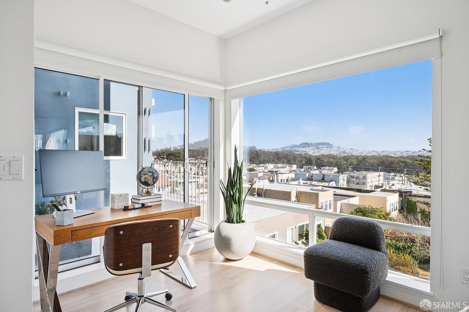 This bright and airy home office features a sleek wooden desk paired with a mid-century modern style chair, positioned to take advantage of the expansive views. Large windows wrap around the corner of the room, flooding the space with natural light and showcasing a panoramic cityscape. A tall potted plant adds a touch of greenery, while the minimalist decor creates a professional yet serene atmosphere.
