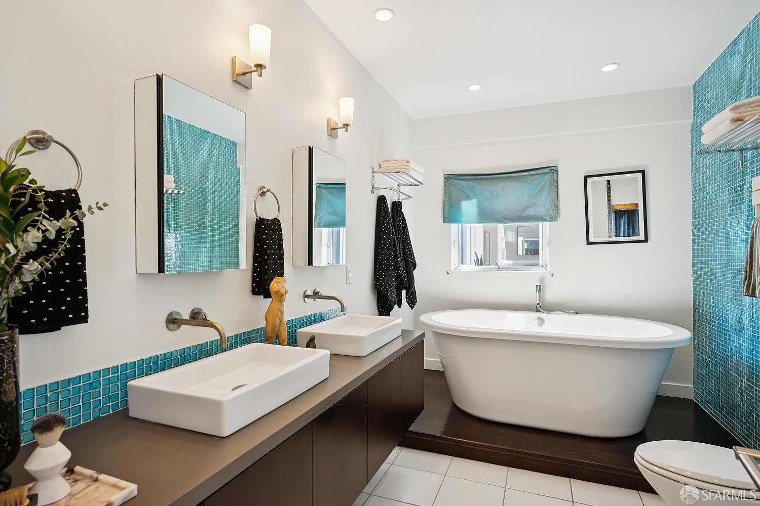 This modern primary bathroom features a dual-sink vanity with rectangular vessel sinks and a striking blue mosaic tile backsplash. A freestanding soaking tub sits on a raised dark wood platform, complemented by a window with a blue shade and a minimalist framed art piece. The space is brightened by recessed lighting and wall-mounted sconces, creating a clean and contemporary aesthetic.