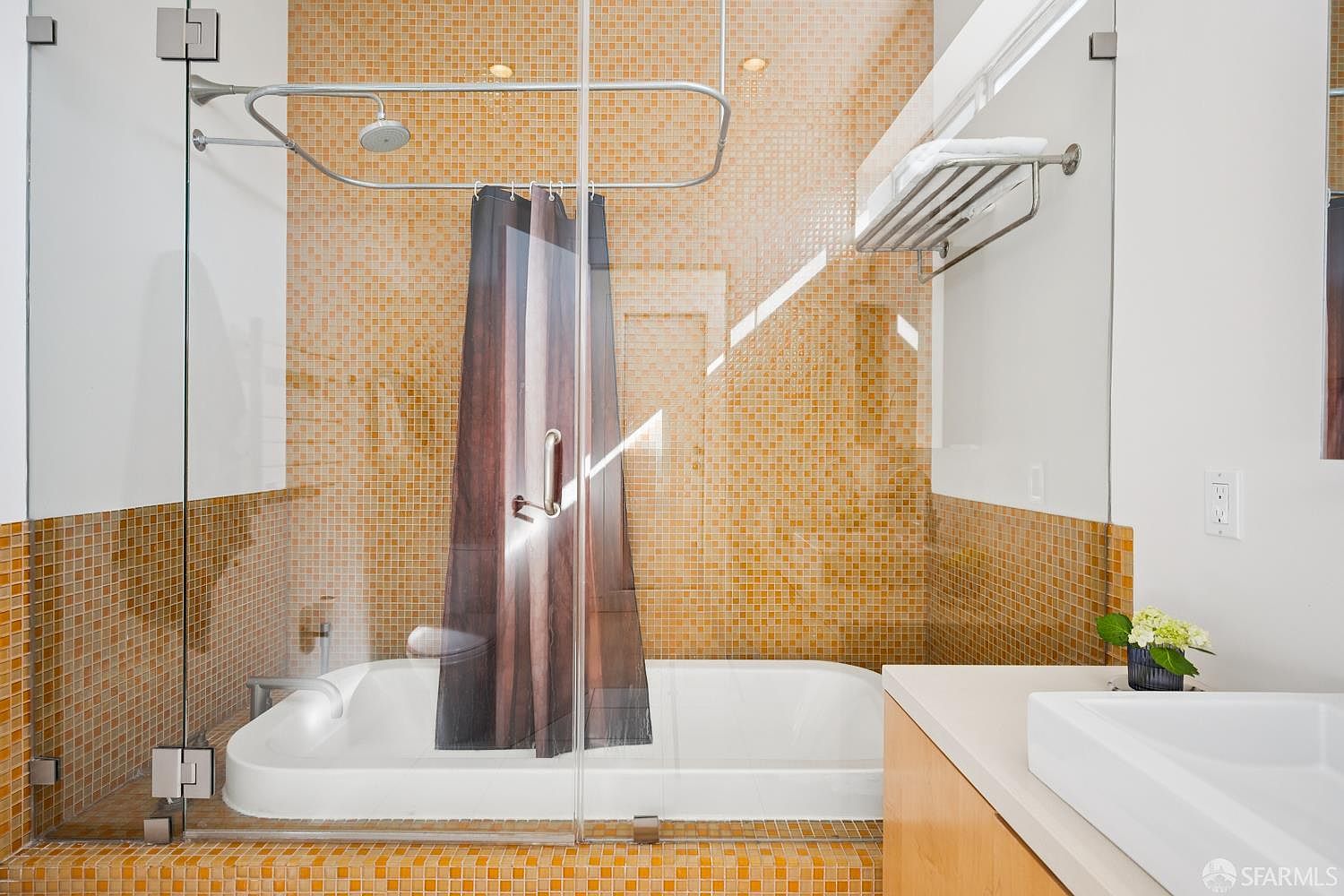 This bathroom features a shower-tub combination enclosed by a glass partition, accented by a dark, patterned shower curtain. The walls are finished with small, square, warm-toned mosaic tiles that create a textured, golden aesthetic. A wall-mounted towel rack is positioned above the glass enclosure, and a portion of a white vanity sink is visible in the foreground.