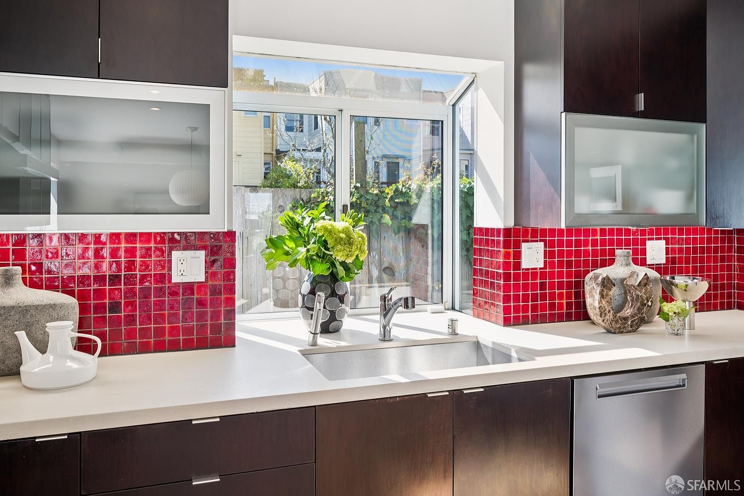 This modern kitchen features dark wood cabinetry contrasted by a vibrant red tiled backsplash and light-colored countertops. A stainless steel sink is positioned beneath a window that looks out onto a private outdoor area with greenery. The space is well-lit with natural light, creating a clean and contemporary aesthetic.