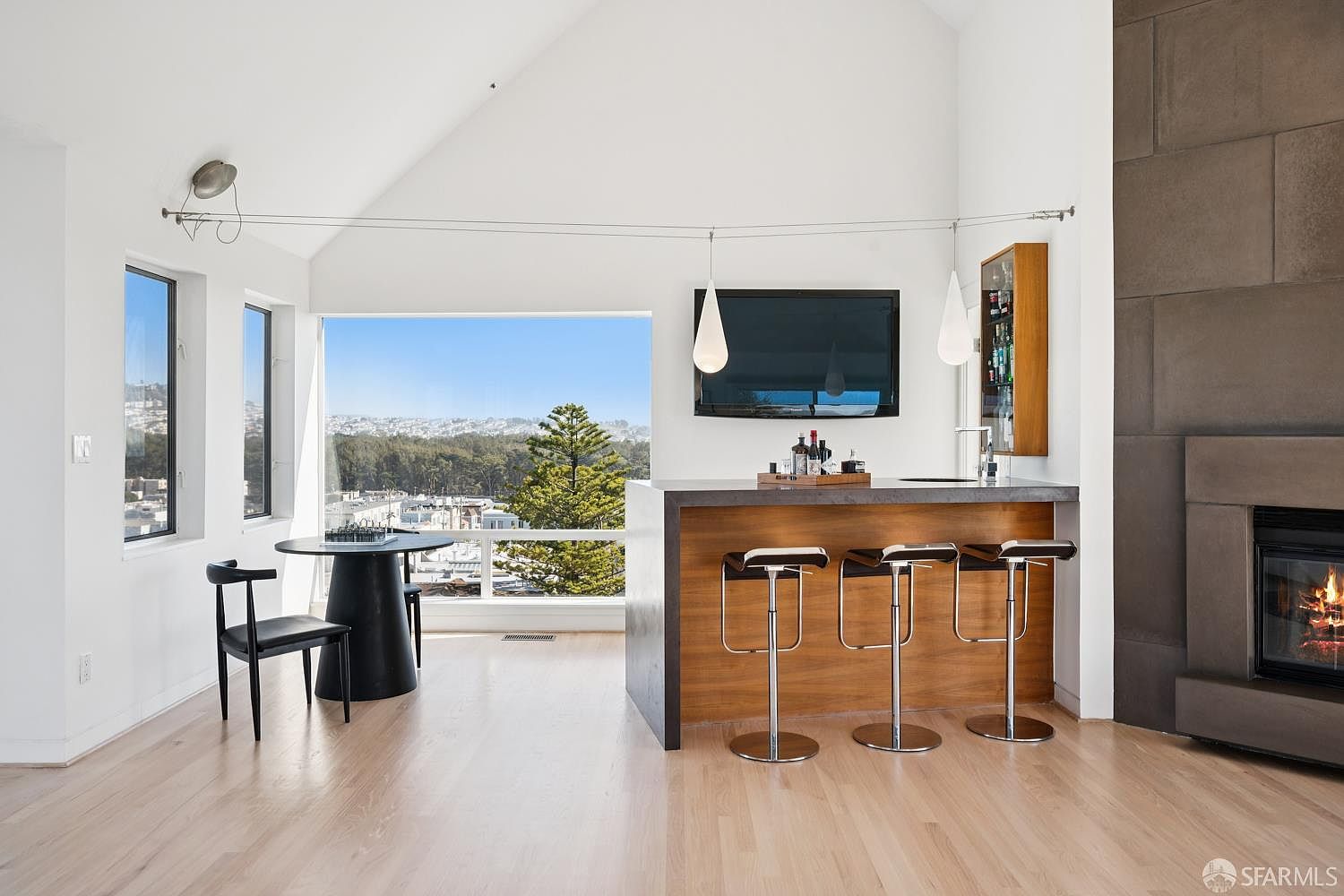 This bright, open-concept living space features a modern bar area with three stools and a sleek, dark-toned counter. To the left, a small dining table sits near large windows that frame a scenic view of the surrounding neighborhood and trees. A tall, dark-toned fireplace surround anchors the right side of the room, creating a sophisticated focal point under the vaulted ceiling.