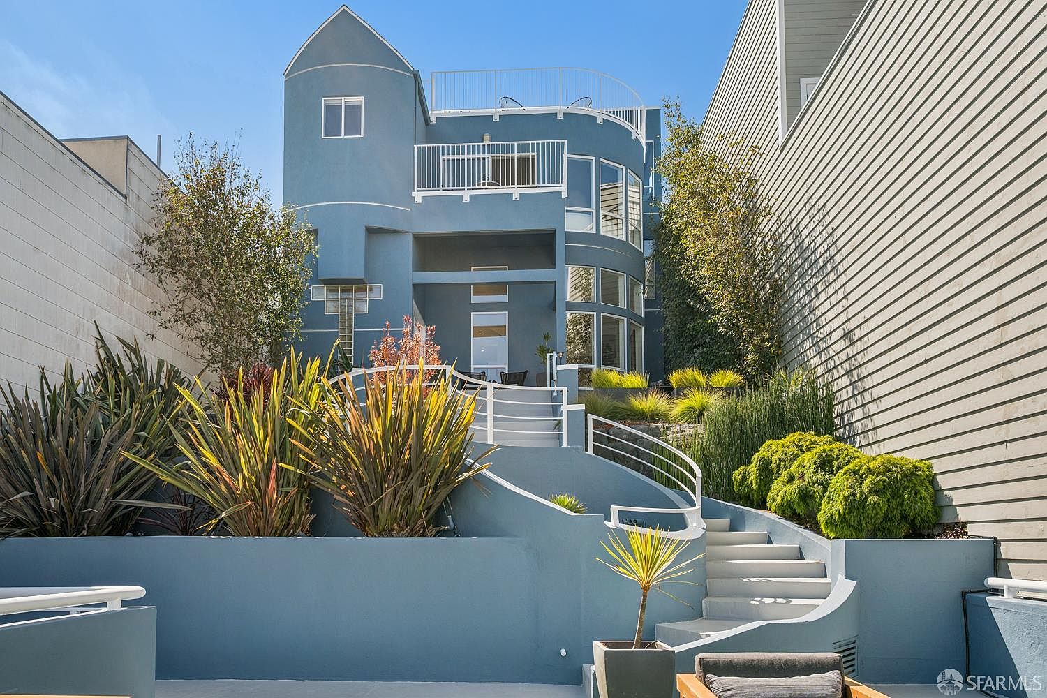 This rear exterior view showcases a multi-level blue home with a distinctive curved architectural design and multiple balconies. A set of curved concrete stairs leads up through a landscaped garden area filled with ornamental grasses and shrubs toward the main entrance. The perspective is from a lower patio level, emphasizing the home's verticality and the tiered transition between the outdoor living space and the structure.
