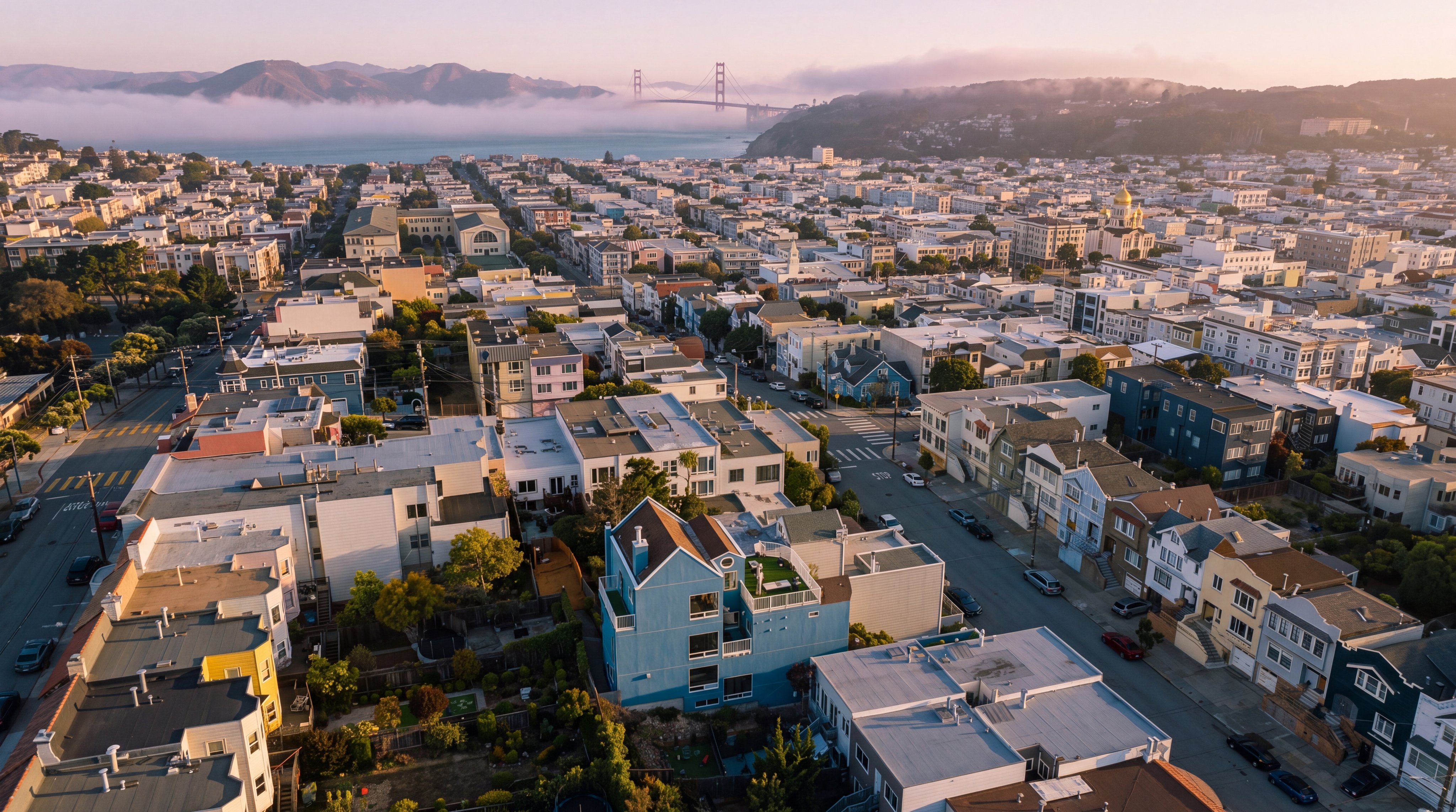 This high-angle aerial view captures a dense urban neighborhood in San Francisco, showcasing a prominent blue multi-story residence with a rooftop deck. The perspective extends across the city grid toward the iconic Golden Gate Bridge visible in the distance against a clear blue sky. The scene highlights the compact, hilly nature of the residential area, characterized by a mix of architectural styles and lush backyard greenery.