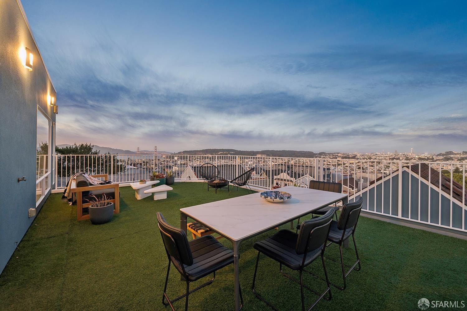 This expansive rooftop deck features artificial turf and a modern dining set, providing an ideal space for outdoor entertaining. The area is enclosed by a white metal railing and offers panoramic views of the city skyline and distant hills under a twilight sky. The setting is complemented by additional lounge seating and a small fire pit, creating a sophisticated and inviting atmosphere.