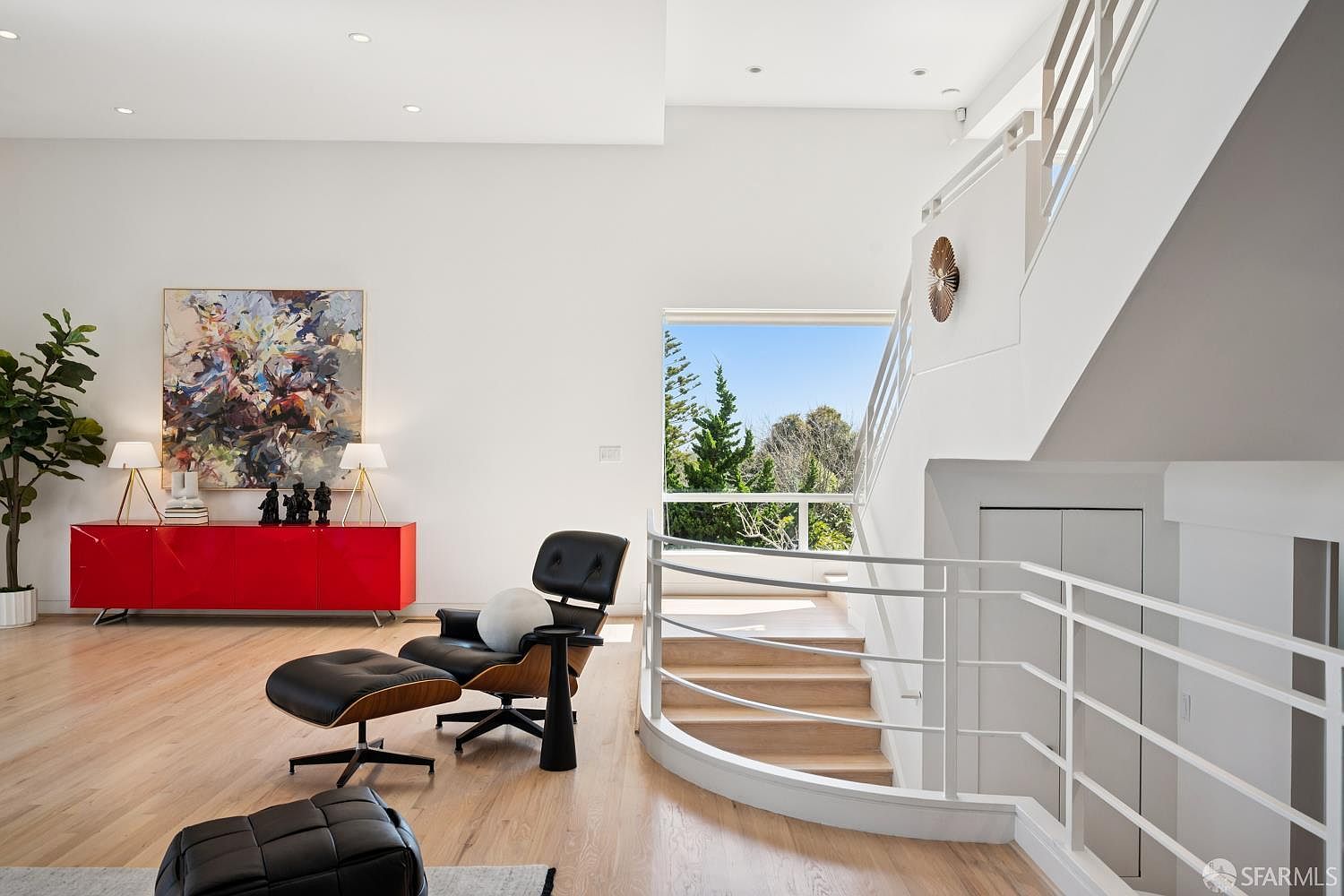 This bright and airy living space features light wood flooring and a minimalist aesthetic, anchored by a striking red console table topped with modern lamps and a large abstract painting. A classic lounge chair with an ottoman sits near a curved staircase with white railings, which leads down to a lower level. Large windows provide a clear view of lush green trees outside, enhancing the open and sophisticated atmosphere of the room.