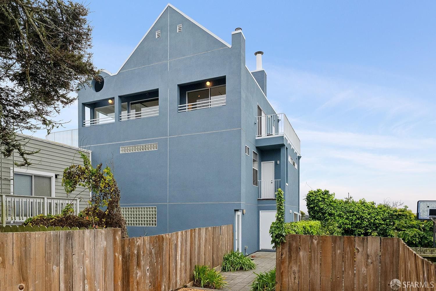 This rear view of a multi-story blue home showcases a contemporary architectural style with a gabled roofline and multiple balconies. The exterior features a mix of smooth siding, a glass block window section, and a small paved patio area enclosed by a wooden fence. The perspective is from ground level, looking up at the structure, which is bordered by mature trees and lush greenery.