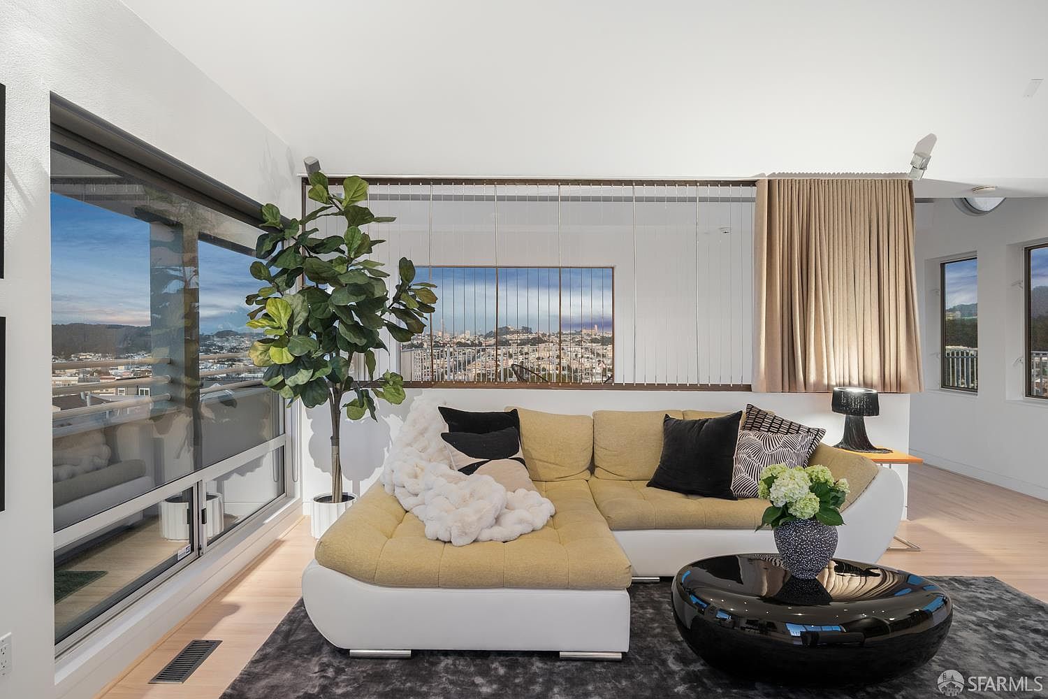 This bright, contemporary living room features a large, L-shaped sectional sofa with a light-colored textured fabric and a white base, accented by a plush white throw and dark decorative pillows. A glossy black circular coffee table sits on a dark gray area rug, while a tall potted plant adds a touch of greenery near the expansive windows that offer a panoramic city view. The space is defined by a unique room divider made of vertical metal cables and a neutral color palette, creating an open and airy atmosphere.