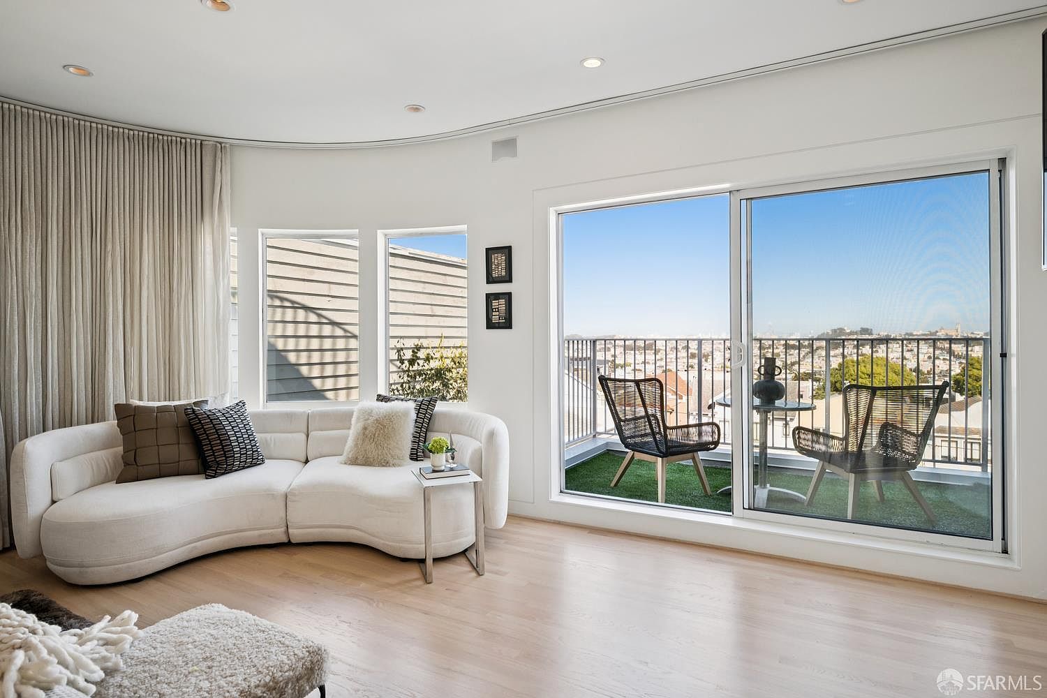 This bright and airy living room features a curved white sofa accented with textured pillows, set against a backdrop of light wood flooring and neutral walls. A large sliding glass door opens onto a private balcony with outdoor seating, offering an expansive view of the city skyline. The space is designed with a modern, minimalist aesthetic, emphasizing natural light and a seamless transition between indoor and outdoor living.