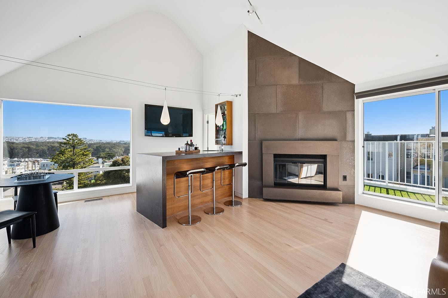 This bright, open-concept living space features a modern bar area with three stools and a prominent fireplace with a tall, dark-toned surround. Large windows on either side of the room provide expansive views and fill the space with natural light, highlighting the light wood flooring. The room is designed with a contemporary aesthetic, blending a functional entertainment area with a sophisticated lounge atmosphere.